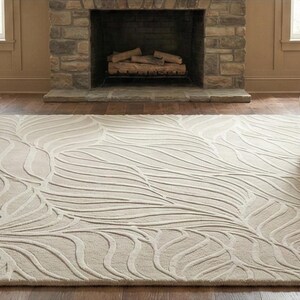 May include: A large, rectangular area rug with a cream-colored background and a raised, leaf-like pattern in a lighter shade. The rug is placed on a hardwood floor, with two armchairs and a fireplace in the background.