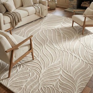 May include: Beige area rug with a leaf-like pattern in a living room setting. The rug features a raised, textured design in a lighter shade of beige, creating a subtle contrast. The room includes a beige sofa, chairs, and a fireplace.