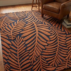 May include: A rectangular area rug with a dark blue background and a repeating pattern of large, raised, rust-colored palm leaves. The rug is placed on a hardwood floor, with a leather armchair visible in the background.