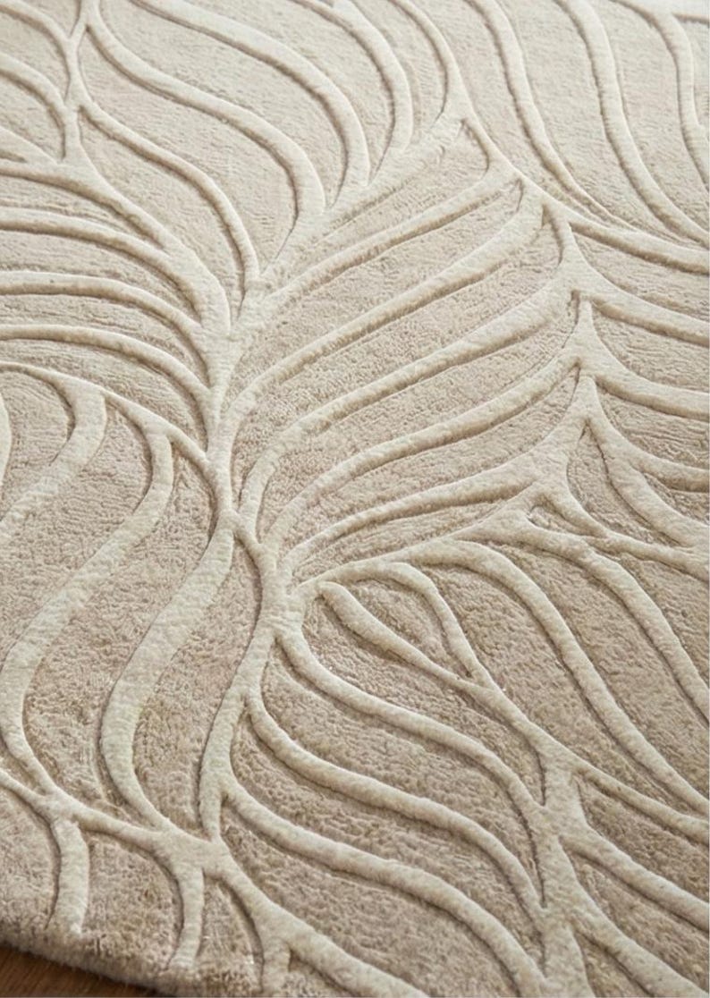 May include: Close-up of a beige rug with a raised leaf pattern. The design features curved, vein-like lines in a lighter shade of beige, creating a textured, three-dimensional effect. The rug's neutral color palette and organic design suggest a natural, modern aesthetic.