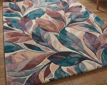 May include: A rectangular area rug with a botanical leaf design. The rug features large, stylized leaves in shades of teal, purple, brown, and beige. The leaves overlap, creating a textured and layered appearance. The rug is placed on a wooden floor.