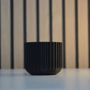 A black, round, ribbed planter pot. The pot has vertical grooves and a rounded base. The background features a light brown wooden wall with vertical dark brown stripes. The pot is sitting on a white surface.