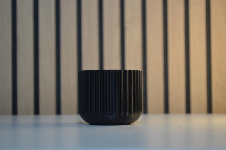 A black, round, ribbed planter pot. The pot has vertical grooves and a rounded base. The background features a light brown wooden wall with vertical dark brown stripes. The pot is sitting on a white surface.