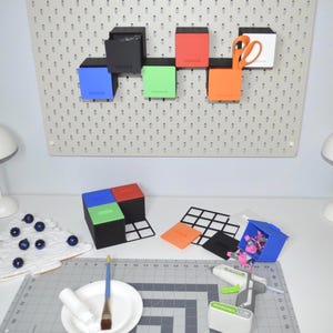 May include: A craft room scene with a pegboard displaying colorful square boxes. Below, a white Christmas tree decoration, paint supplies, and a glue gun are arranged on a cutting mat. The boxes are in various colors: black, red, orange, green, blue, and white.