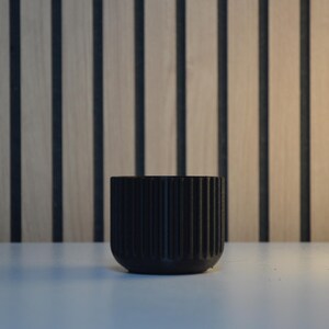 A black, cylindrical planter with vertical ribbed detailing. The small, modern pot is made of a matte material and sits on a white surface. The background features a wooden wall with vertical black stripes.