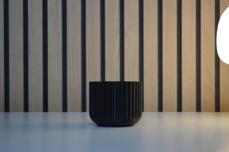 A black, cylindrical planter with vertical ribbed detailing. The small, modern pot is made of a matte material and sits on a white surface. The background features a wooden wall with vertical black stripes.