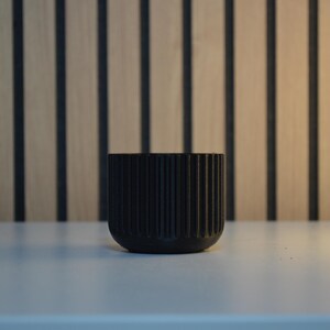 A black, ribbed ceramic planter with a rounded base. The planter is cylindrical and features vertical grooves. The background has a wooden panel design with vertical black stripes. The planter is sitting on a white surface.