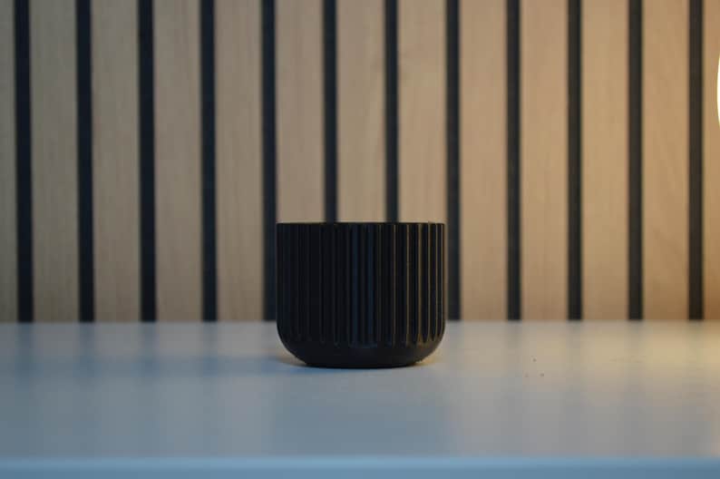 A black, ribbed ceramic planter with a rounded base. The planter is cylindrical and features vertical grooves. The background has a wooden panel design with vertical black stripes. The planter is sitting on a white surface.