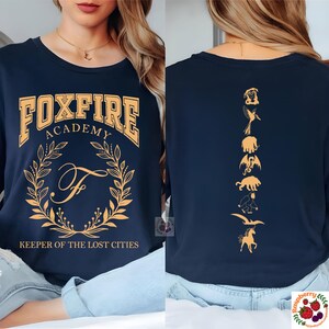 Op de afbeelding: Marineblauwe sweatshirt met lange mouwen met "FOXFIRE ACADEMY" in gouden letters. De voorkant heeft een gouden krans en de letter "F". De achterkant toont gouden illustraties van mythische wezens. "KEEPER OF THE LOST CITIES" staat eronder.