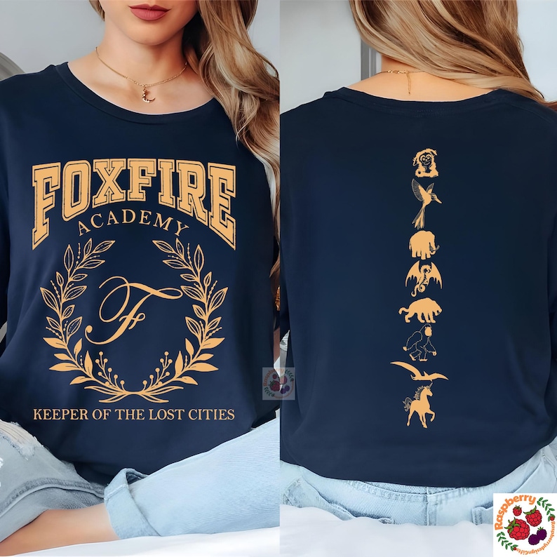 Op de afbeelding: Marineblauwe sweatshirt met lange mouwen met "FOXFIRE ACADEMY" in gouden letters. De voorkant heeft een gouden krans en de letter "F". De achterkant toont gouden illustraties van mythische wezens. "KEEPER OF THE LOST CITIES" staat eronder.