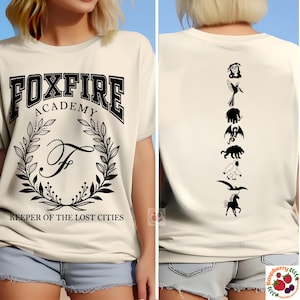Op de afbeelding: Cr&egrave;mekleurig T-shirt met "FOXFIRE ACADEMY" tekst en een lauwerkrans ontwerp. De achterkant heeft een verticale rij zwarte silhouet fantasie wezens. Tekst "KEEPER OF THE LOST CITIES" eronder.
