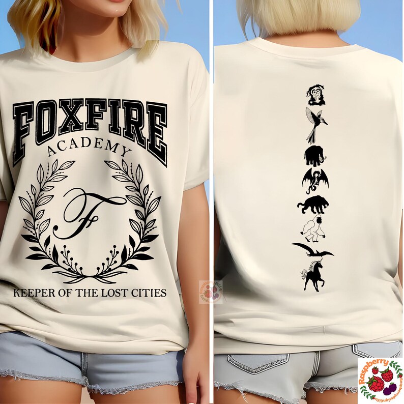 Op de afbeelding: Cr&egrave;mekleurig T-shirt met "FOXFIRE ACADEMY" tekst en een lauwerkrans ontwerp. De achterkant heeft een verticale rij zwarte silhouet fantasie wezens. Tekst "KEEPER OF THE LOST CITIES" eronder.