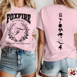 Op de afbeelding: Lichtroze t-shirt met de zwarte tekst "FOXFIRE ACADEMY" boven een kransontwerp en de tekst "KEEPER OF THE LOST CITIES". De achterkant van het shirt toont zwarte silhouetten van wezens.