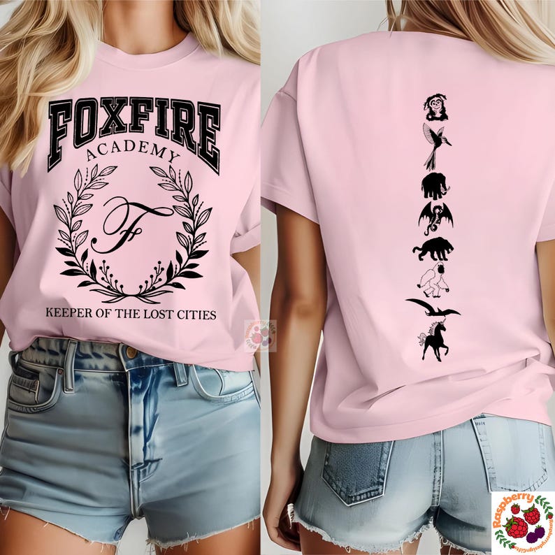 Op de afbeelding: Lichtroze t-shirt met de zwarte tekst "FOXFIRE ACADEMY" boven een kransontwerp en de tekst "KEEPER OF THE LOST CITIES". De achterkant van het shirt toont zwarte silhouetten van wezens.