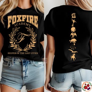 Op de afbeelding: Zwart T-shirt met de tekst "FOXFIRE ACADEMY" en een gouden kransontwerp. De achterkant van het shirt heeft een verticale lijn van gouden mythische wezens. "KEEPER OF THE LOST CITIES" staat eronder.