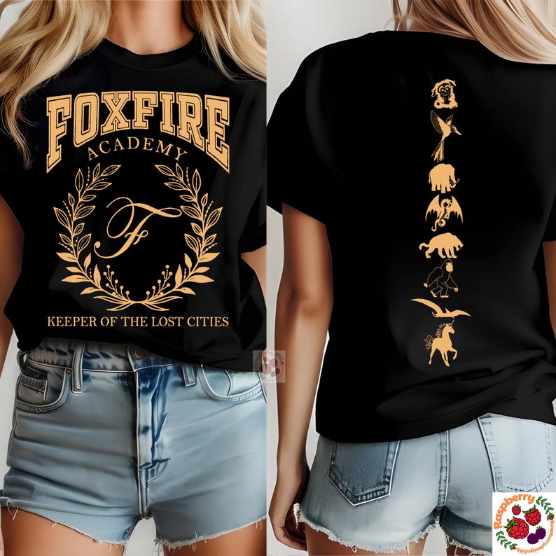 Op de afbeelding: Zwart T-shirt met de tekst "FOXFIRE ACADEMY" en een gouden kransontwerp. De achterkant van het shirt heeft een verticale lijn van gouden mythische wezens. "KEEPER OF THE LOST CITIES" staat eronder.