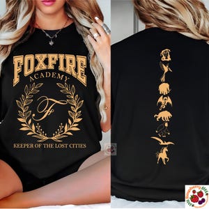 Op de afbeelding: Zwart T-shirt met gouden "FOXFIRE ACADEMY" tekst, een lauwerkrans en de letter "F". De achterkant toont gouden silhouetten van mythische wezens. "KEEPER OF THE LOST CITIES" is eronder gedrukt.