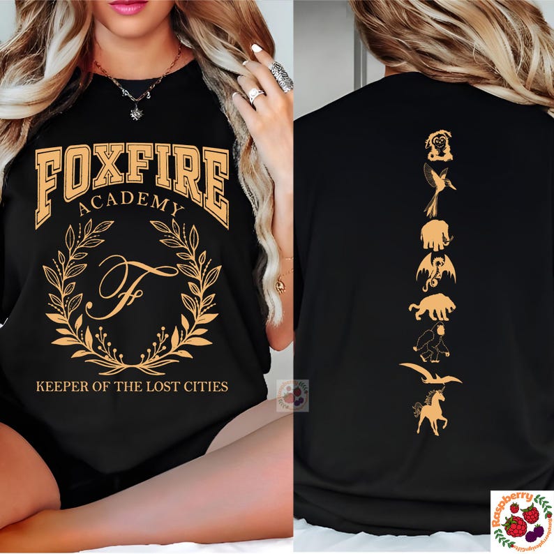 Op de afbeelding: Zwart T-shirt met gouden "FOXFIRE ACADEMY" tekst, een lauwerkrans en de letter "F". De achterkant toont gouden silhouetten van mythische wezens. "KEEPER OF THE LOST CITIES" is eronder gedrukt.