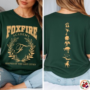 Op de afbeelding: Donkergroen t-shirt met gouden "FOXFIRE ACADEMY" letters. De voorkant heeft een lauwerkrans en de letter "F". De achterkant toont gouden silhouetten van mythische wezens. "KEEPER OF THE LOST CITIES" staat eronder.