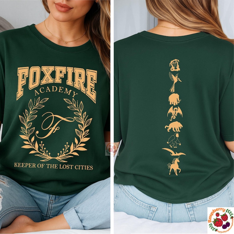 Op de afbeelding: Donkergroen t-shirt met gouden "FOXFIRE ACADEMY" letters. De voorkant heeft een lauwerkrans en de letter "F". De achterkant toont gouden silhouetten van mythische wezens. "KEEPER OF THE LOST CITIES" staat eronder.