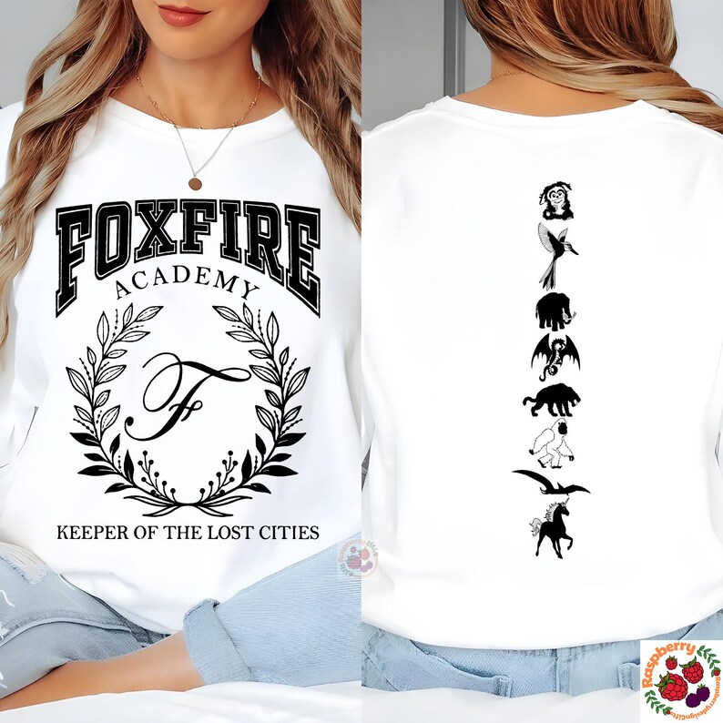 Op de afbeelding: Wit sweatshirt met de tekst "FOXFIRE ACADEMY" in een collegiale letter, een lauwerkrans en de letter "F". De achterkant toont zwarte silhouetten van wezens. De tekst "KEEPER OF THE LOST CITIES" staat eronder.