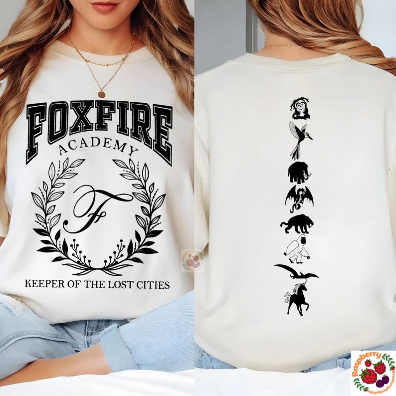 Op de afbeelding: Cr&egrave;mekleurige sweatshirt met de tekst "FOXFIRE ACADEMY" en een lauwerkransontwerp. De voorkant heeft de tekst "KEEPER OF THE LOST CITIES". De achterkant heeft een verticale rij zwarte fantasiefiguren.