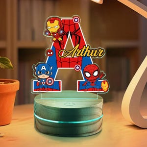 Puede incluir: Una luz nocturna de acrílico personalizada con una letra A grande con un patrón de telaraña rojo y azul. El diseño incluye ilustraciones de estilo caricatura de Iron Man, Capitán América y Spider-Man, con el nombre Arthur en letra dorada.