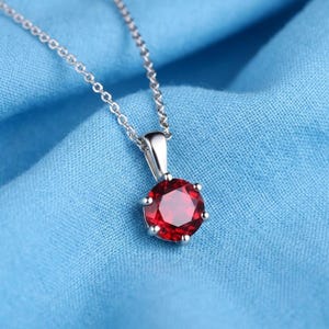 May include: A silver-toned necklace featuring a round, faceted, deep red gemstone pendant held in a six-prong setting. The pendant is suspended from a delicate, thin silver-toned chain that connects to the top of the metal bail.