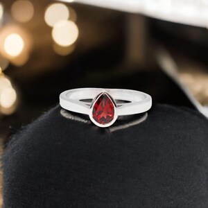 May include: A silver ring with a pear-shaped, deep red gemstone. The ring's band is a simple, polished silver. The gemstone is set in a bezel setting, and the ring is displayed on a black surface.