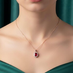 May include: A rose gold necklace with a pendant. The pendant has a large oval, deep red gemstone, encircled by smaller, clear gemstones. The necklace is shown against a green backdrop.