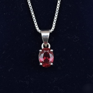 Op de afbeelding: Een zilveren hangerketting met een ovaalvormige, dieprode edelsteen. De edelsteen is gezet in een zilveren zetting en hangt aan een fijne zilveren ketting. De achtergrond is donkerblauw.