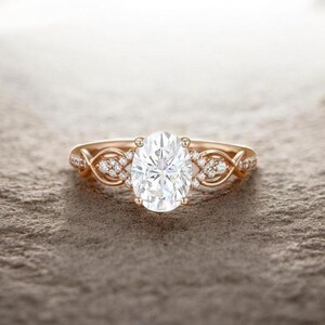 May include: Rose gold engagement ring featuring an oval-cut diamond center stone. The band has a braided design with small accent diamonds. The ring is set on a textured surface, showcasing the intricate details of the jewelry.