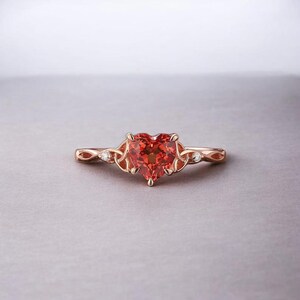 Pode incluir: Anel de ouro rosa com uma pedra preciosa vermelho-alaranjada em forma de coração. O anel tem uma banda delicada e torcida e pequenas pedras de destaque. A cravação apresenta um design de nó celta.