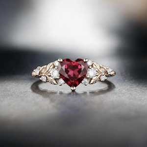 Może przedstawiać: Pierścionek z różowego złota z centralnie umieszczonym, głębokim, czerwonym kamieniem szlachetnym w kształcie serca. Pierścionek zdobiony jest małymi, okrągłymi, przezroczystymi kamieniami szlachetnymi i detalami w kształcie liści na obręczy. Pierścionek leży na odblaskowej powierzchni.