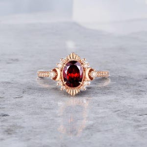 Może przedstawiać: Pierścionek z różowego złota z owalnym, głęboko czerwonym kamieniem szlachetnym. Pierścionek ma szczegółową oprawę z motywem promieni słonecznych wokół głównego kamienia, zaakcentowaną mniejszymi białymi kamieniami szlachetnymi i kształtami półksiężyca.