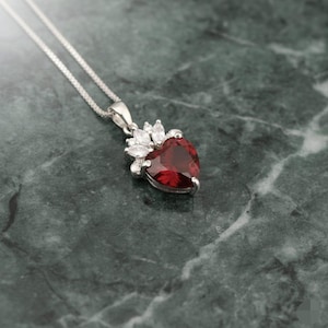 May include: A silver necklace featuring a heart-shaped, deep red pendant. The pendant is framed by smaller, white gemstones. The necklace is shown on a dark green, marble-effect surface.