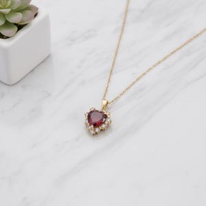 May include: A gold necklace with a heart-shaped pendant. The pendant has a large, deep red gemstone surrounded by smaller, clear gemstones. The necklace is displayed on a white marble surface.