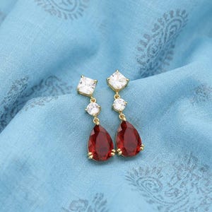 Peut inclure: Une paire de boucles d'oreilles pendantes dorées avec un design carré, rond et en forme de goutte. Les boucles d'oreilles présentent des pierres carrées et rondes transparentes, et des pierres en forme de goutte rouge foncé. Sur un tissu bleu.