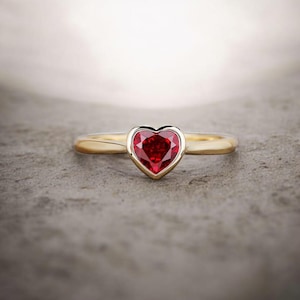 Może przedstawiać: Złoty pierścionek z sercowatym, głęboko czerwonym kamieniem szlachetnym. Kamień jest osadzony w oprawie z ramką, a obrączka jest gładka i wypolerowana. Pierścionek jest prezentowany na teksturowanej, szarej powierzchni.