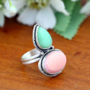 May include: A silver ring featuring two distinct gemstones set in twisted metal frames. The top stone is a light green teardrop shape, while the bottom stone is a smooth, oval-shaped pink cabochon. The band is made of a simple, polished silver metal.