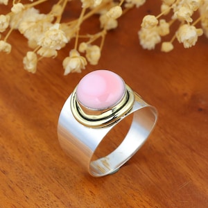 May include: A wide band ring featuring a smooth, round, light pink cabochon stone set in a gold-toned bezel. The ring band is made of polished silver-toned metal, creating a contrast between the gold-toned setting and the silver-toned band.