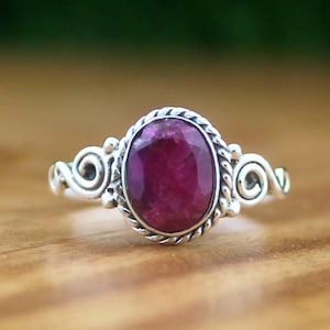 Op de afbeelding: Een zilveren ring met een ovale, dieprode edelsteen. De edelsteen is gezet in een gedraaide, touwachtige bezel, met decoratieve krullen aan weerszijden. De ring wordt gepresenteerd op een houten oppervlak.