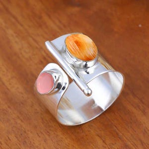 Puede incluir: Un anillo de banda ancha de plata con un diseño abierto y una barra horizontal. El anillo está adornado con dos piedras engastadas: una piedra más grande de forma ovalada con rayas naranjas y blancas y una piedra redonda más pequeña de color rosa claro.