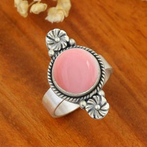 May include: A silver ring featuring a smooth, round, light pink cabochon stone set in a twisted rope border. The band is accented with two small, circular, flower-shaped silver motifs positioned at the top and bottom of the central stone setting.