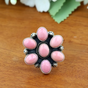 May include: A silver ring featuring a central oval pink stone surrounded by six smaller oval pink stones arranged in a floral pattern. The setting includes a twisted rope border around the centre stone and small silver starburst accents.