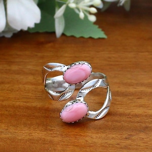 Könnte beinhalten: Ein silberner Ring mit zwei ovalen, hellrosa Edelsteinen. Der Ring hat ein offenes Design mit geätzten Details. Die Edelsteine sind in einer Zargenfassung gefasst. Der Ring wird auf einer Holzoberfläche präsentiert.