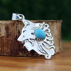 May include: A silver pendant featuring a wolf's head design with a turquoise stone center. The pendant also includes a profile of a woman and decorative flourishes. The pendant is displayed on a wooden surface.
