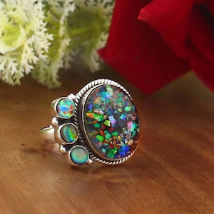 Puede incluir: Anillo de plata con una gran piedra de ópalo ovalada. El ópalo presenta un mosaico de colores que incluyen verde, azul, rojo y amarillo. Tres ópalos más pequeños están engastados en el lateral del anillo. El anillo está sobre una superficie de madera.