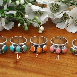 May include: Five silver rings with open bands, each featuring three teardrop-shaped stones in turquoise, orange, pink, and green. The rings are labelled a, b, c, d, and e, showcasing varied stone colours set in textured silver metal bands.