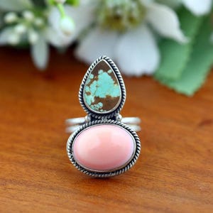May include: A silver ring featuring two stacked stones set in twisted metal wire. The top stone is a teardrop-shaped turquoise with brown matrix, and the bottom stone is a smooth, oval-shaped light pink cabochon. The band is made of silver.